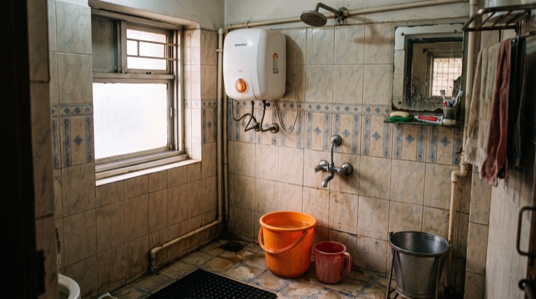 Warm Indian bathroom in morning light, wall-mounted geyser with a small copper indicator glowing, middle-class Kolkata apartment