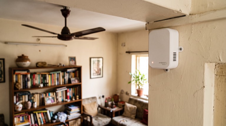 Compact white wall-mounted sensor, about 60 by 40 millimetres, in the corner of a lived-in living room with a faint copper LED