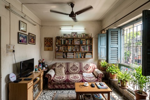 Living room of a middle-class Kolkata apartment in morning light, ceiling fan, bookshelf, lived-in and warm